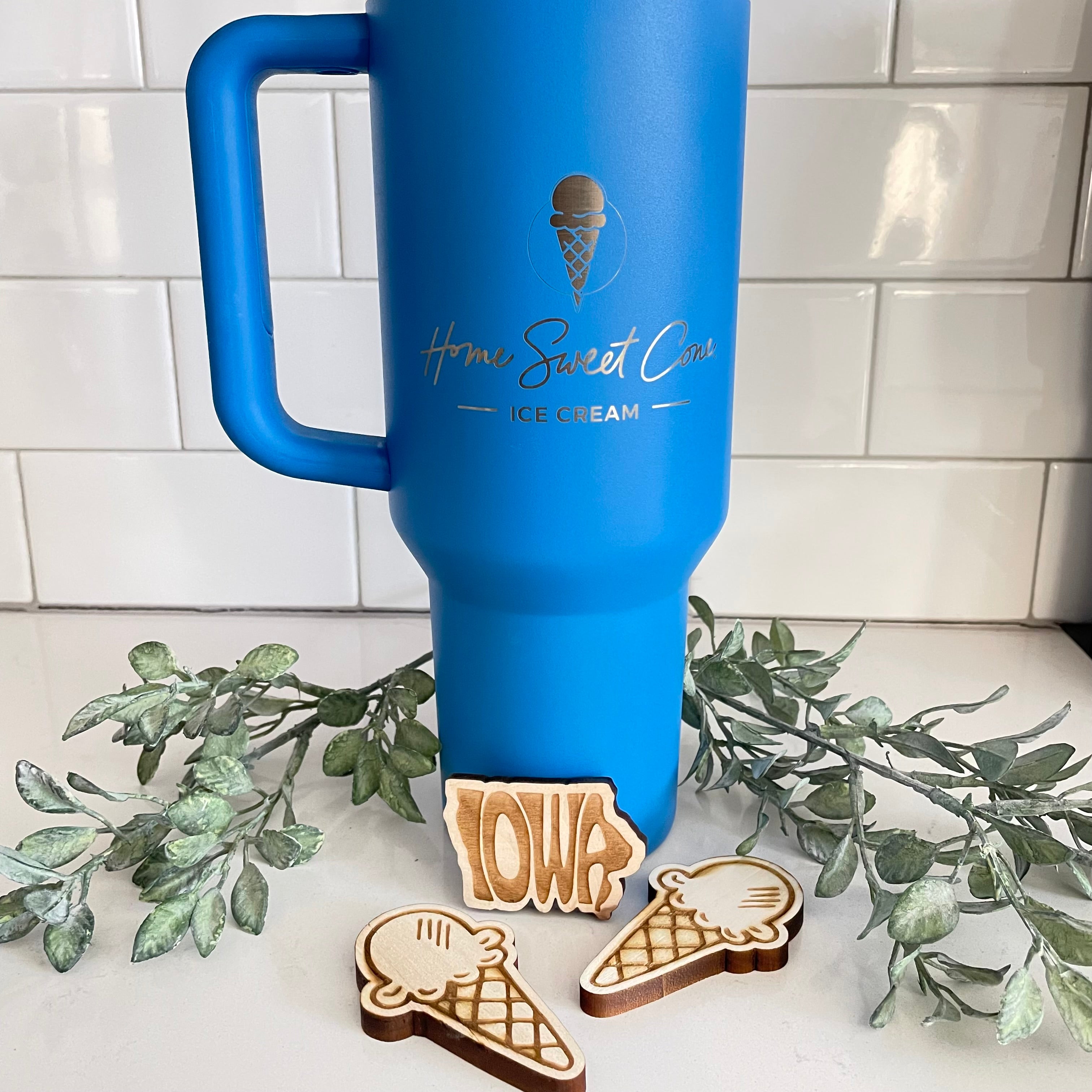 Blue tumbler with Home Sweet Cone engraving and wooden ice cream magnets on a countertop with greenery decor.